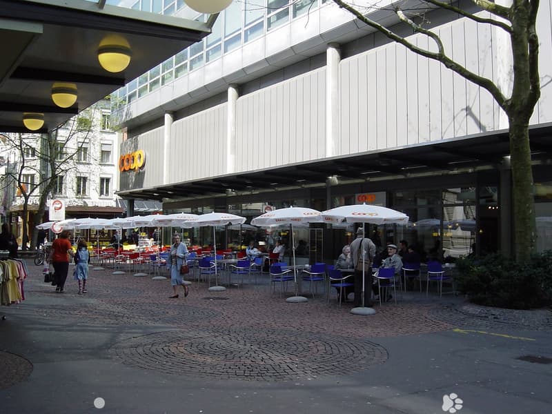 An outdoor seating area with tables and chairs under white umbrellas at a restaurant, surrounded by plants and trees with a brick floor, beside a large building with signage and an open entrance