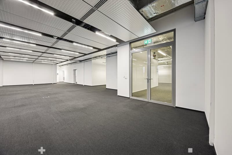 spacious room with gray flooring, white walls, glass doors and windows, ceiling lights, exit sign