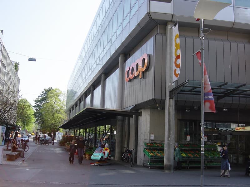 Gray building with the words 'COOP' on it, has awning, bicycles parked in front, person sitting on bench, several people walking on the street, fruit baskets on the side, and street lights