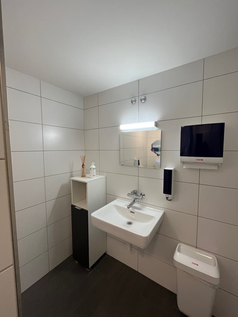 The bathroom has white tiled walls, a white sink with a faucet, and a wall-mounted soap dispenser. It also features a mirror, a toilet paper dispenser, and a small shelf with a bottle, a cup, and a small glass jar. The room has black tile flooring and rec
