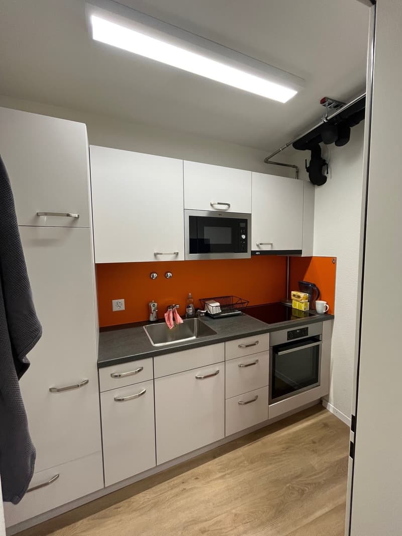 Modern kitchen with white cabinets, orange wall, integrated microwave, sink, and oven. Stainless steel countertop and wooden floor. A towel is hanging on the left side, and a power outlet and faucet are on the wall.