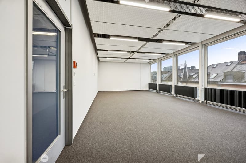 An empty room with carpet flooring, white walls, and glass windows with large radiators beneath. It also has a white door.
