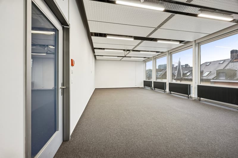 An empty room with carpet flooring, white walls, and glass windows with large radiators beneath. It also has a white door.