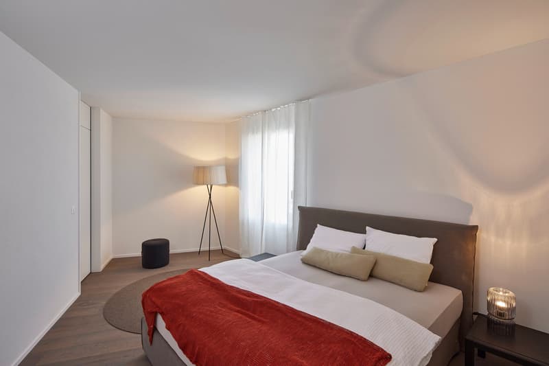 Spacious bedroom with a large bed, white bedding, and a red throw blanket. The room has a floor lamp, a side table, and a curtained window, suggesting a balcony or terrace area.