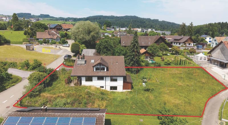 House with brown roof, white walls, solar panels on the roof, surrounded by a large grassy area, located in a rural area with other houses and mountains in the background.