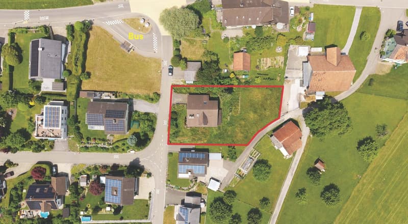 House with solar panels, surrounded by green grass, driveway, and a bus stop nearby