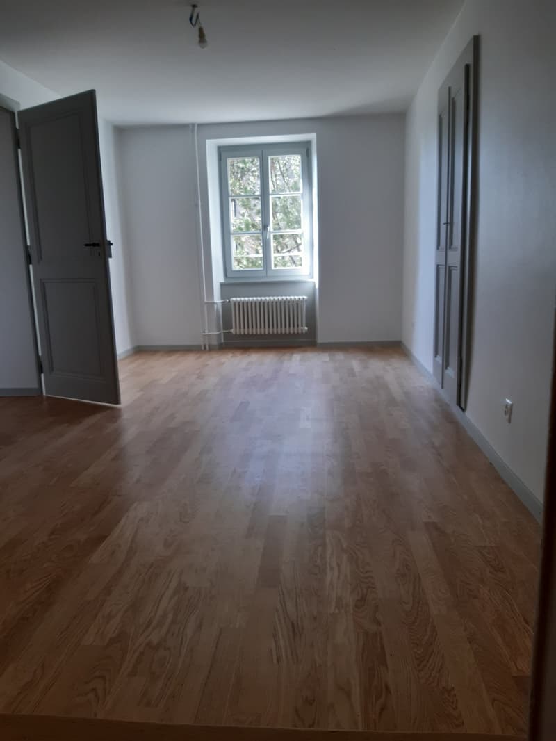 Empty room with wooden floors, white walls, radiator under window, open door, closed door.
