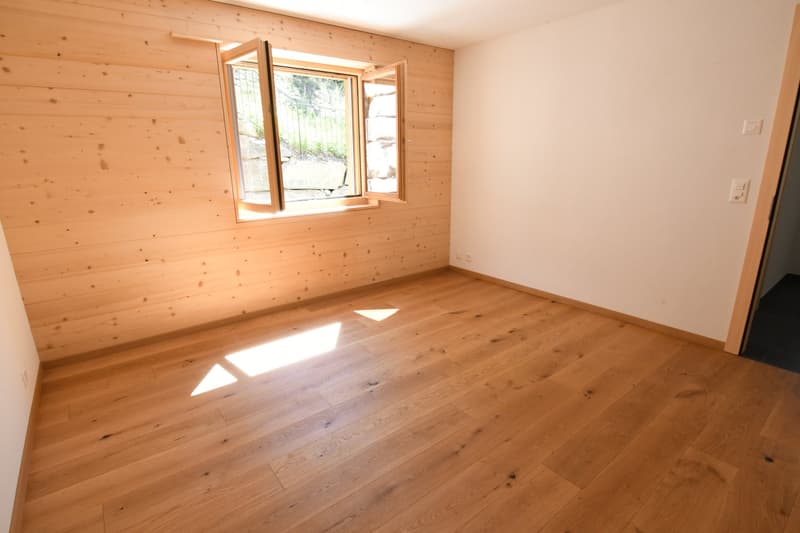 Empty room with wooden floor and walls, two windows with view of outside, one wall painted white