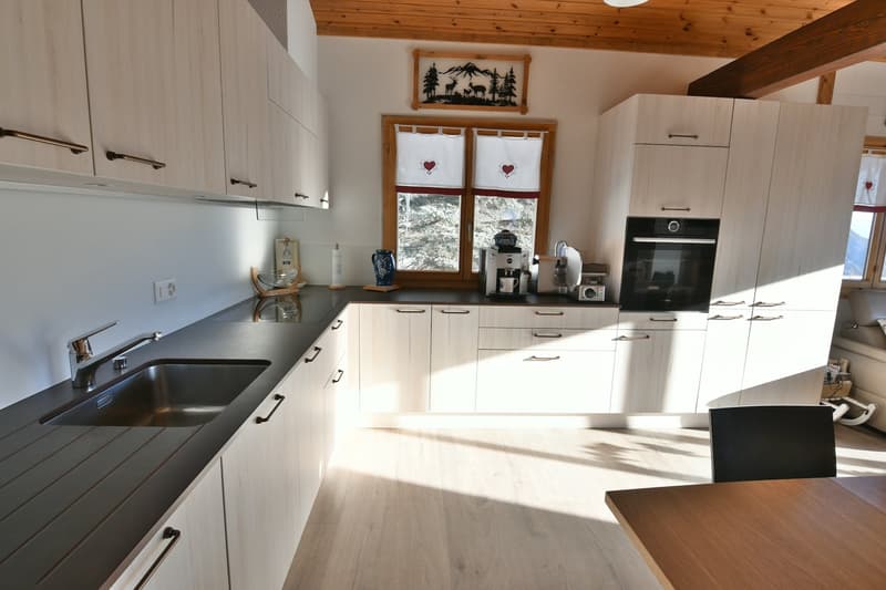 modern kitchen, white cabinets, black countertops, wooden table, stainless steel sink, under-mount sink, dishwasher, coffee machine, built-in oven, window with red and white curtains, wooden ceiling
