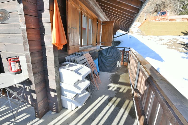 balcony with a wooden railing, wooden wall, stack of plastic chairs, covered grill, outdoor table, and lantern on top of the table