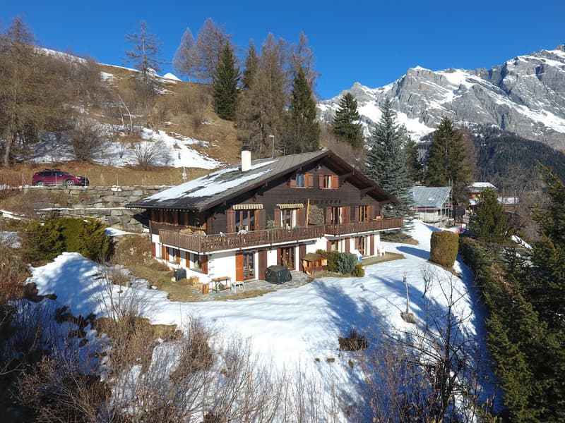 Two-story chalet, wooden exterior, snow on roof, balcony on second floor, snow-covered surroundings, red car parked nearby, surrounded by trees and mountains in the background