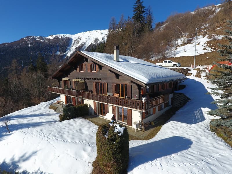 Two-story chalet, wooden exterior, snow-covered roof, large balcony with railing, multiple windows, chimney, driveway leading to the house, car parked nearby, surrounded by snow and trees, mountains in the background