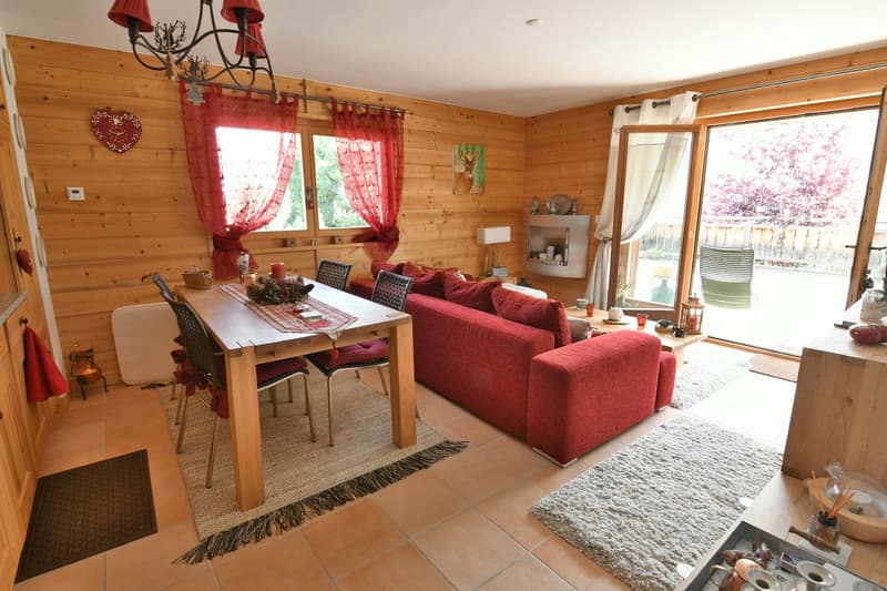 Cozy living room with wooden walls, red curtains, a large red sofa, a wooden dining table, and a view of the outdoors through the sliding glass door.
