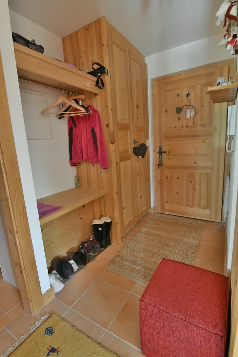 Wooden entryway with storage cabinets, coat hangers, and a red upholstered bench. Tile flooring and a rug in the entrance area.