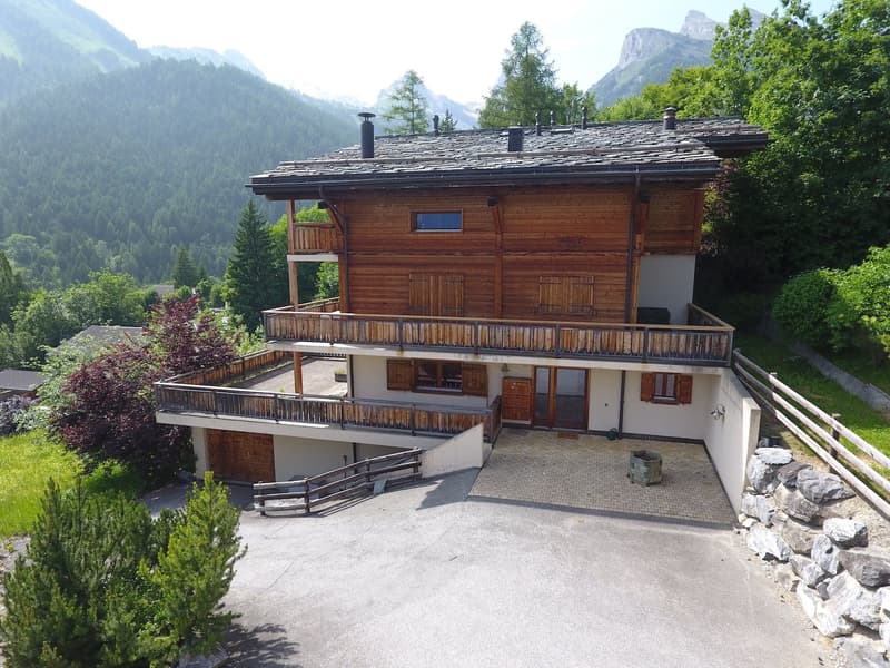 2-story wooden chalet-style house with a balcony, surrounded by lush greenery and mountains in the background