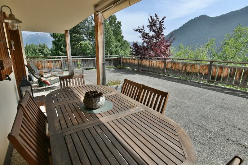 Covered wooden patio with a dining table and chairs, overlooking a scenic mountain landscape with trees and greenery.