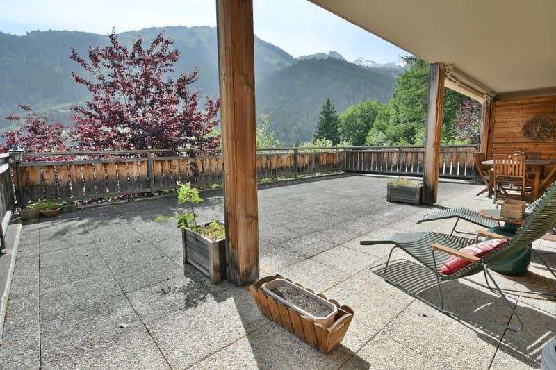Wooden balcony or terrace with a scenic view of the mountains and forests. The balcony has a concrete floor, wooden railings, and some potted plants. There are chairs and other outdoor furniture on the balcony, suggesting it is a relaxing outdoor living s