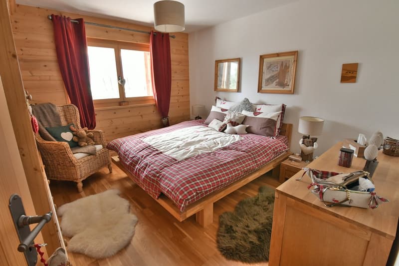 Cozy bedroom with wooden walls, large bed with plaid bedding, wicker chair, framed artwork, and fur rugs on the hardwood floor.