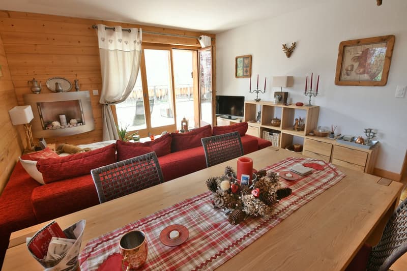 Cozy living room with wooden walls, large windows, red sofa, wooden shelves and cabinets, decorative items like candles and pine cones, and a plaid tablecloth on the coffee table.