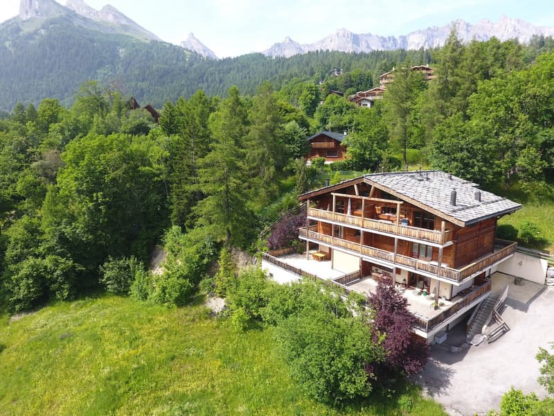 Large wooden chalet-style house with multiple balconies and terraces, surrounded by lush green forest and mountains in the background.