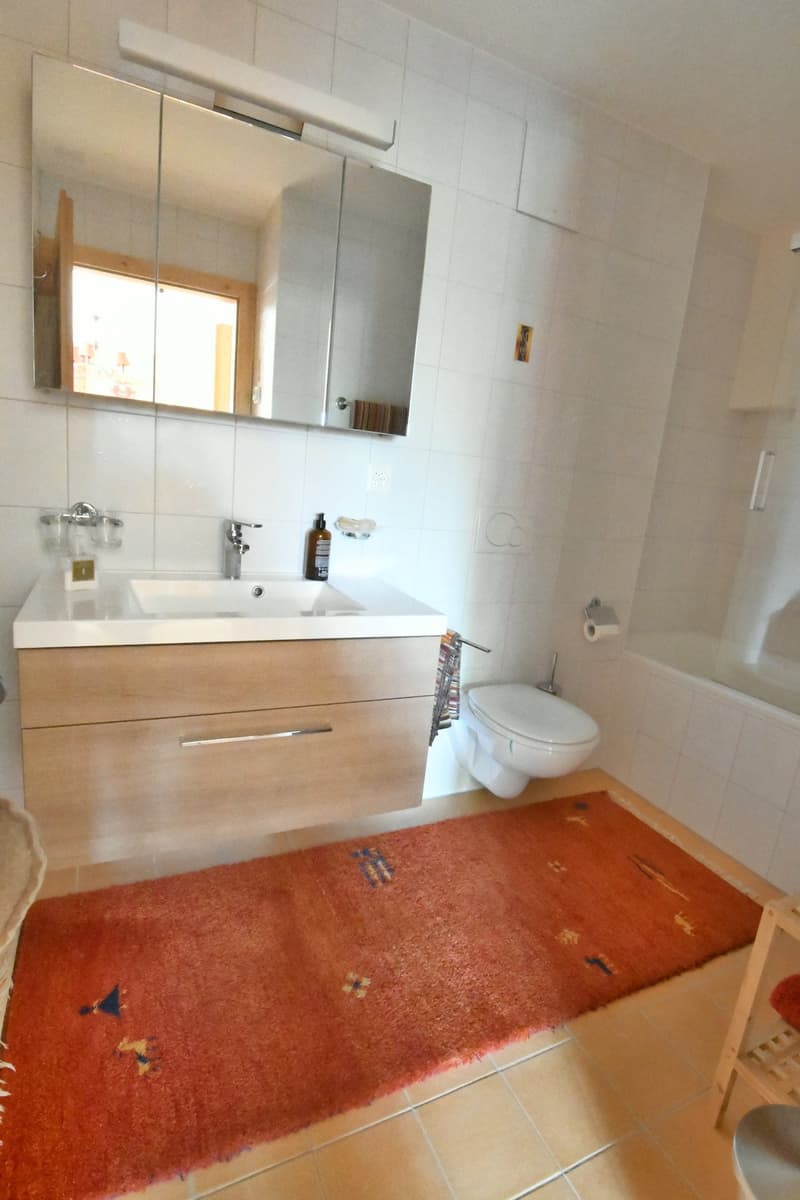 Bathroom with white tiles, vanity with sink, mirror cabinet, and a toilet. There is a red patterned rug on the floor.