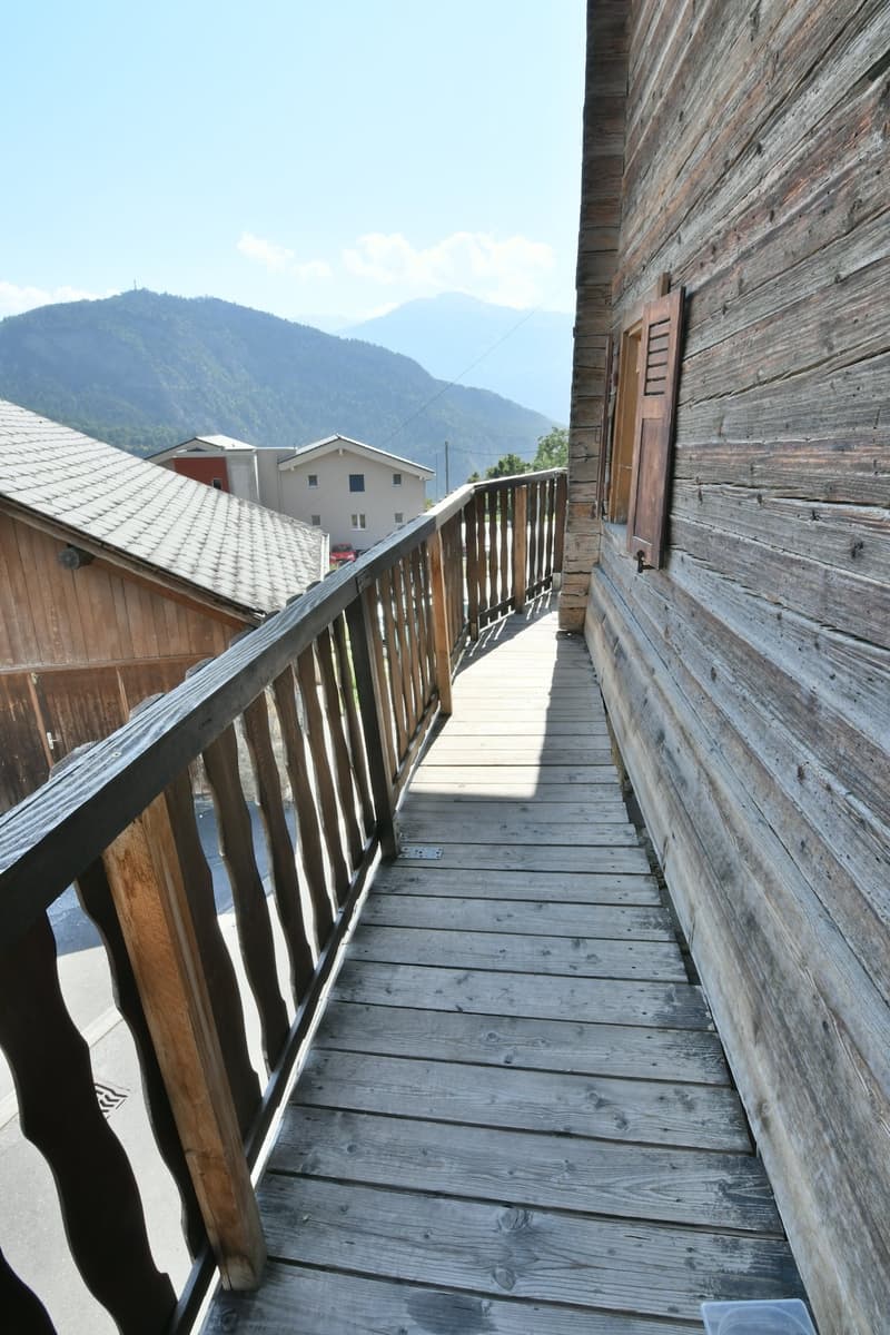 A wooden balcony with a railing, wooden floor, wooden wall, with a view of a mountain and houses. Trees and a road are in the background.