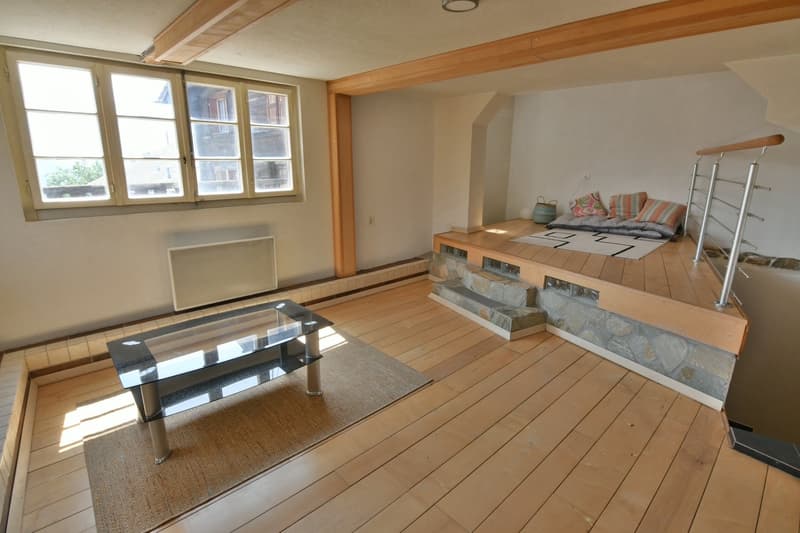 Open space, wooden floor, natural light from window, cozy seating area with pillows, coffee table, radiator on wall, wooden handrail