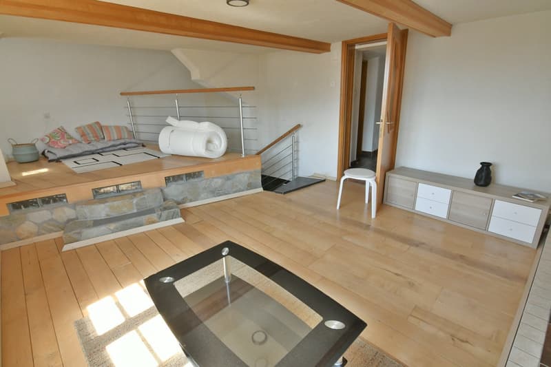 open plan living/sleeping area, hardwood floors, living room sofa with cushions, bed with bedding, white walls, kitchen with counter, window, balcony with railing, glass door to stairs, stool, wooden cabinet with drawers, and a black vase