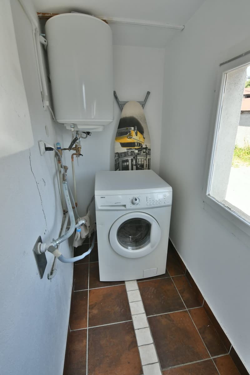 A small room with a white washer machine in the corner, tiled floor, white walls, a window on the right, and an iron rod mounted on the wall with a surfboard.