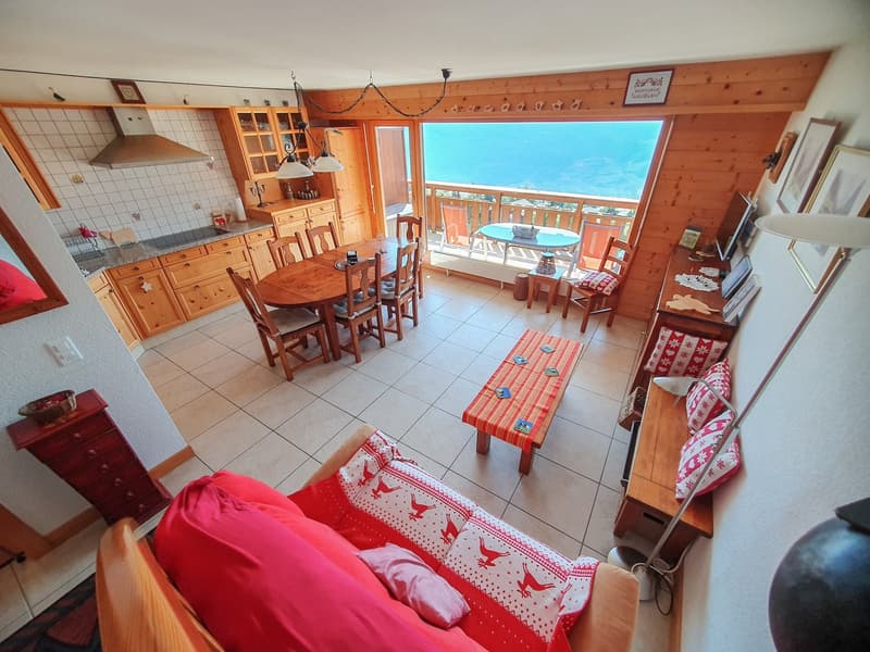 Open living room with wooden dining table, living room couch, and balcony with view. Wooden cabinets, tiled flooring, decorative items, and lamps are present. Kitchen area with stove and microwave. Dining table set with chairs. Balcony has table, chairs,