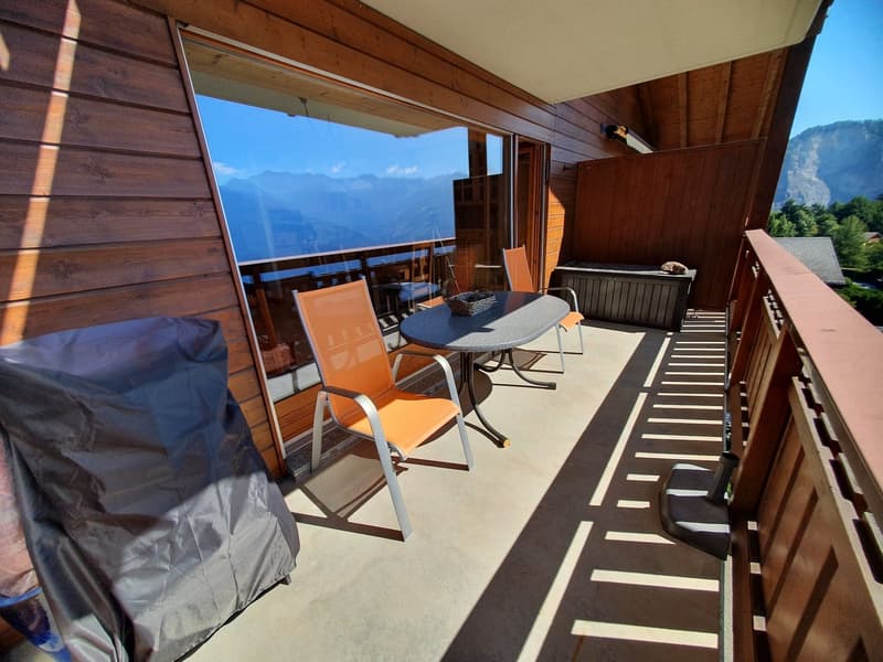 Outdoor space with wooden walls and flooring, outdoor table and chairs, covered area, table has basket on it, covered grill next to the wall, railing, covered storage box, mountain view through the glass windows.