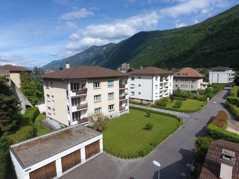 Multi-story apartment building with brown tiled roofs, surrounded by green lawns and gardens, with balconies visible on some units. The building is situated in a mountainous, forested area with a clear blue sky and clouds overhead.
