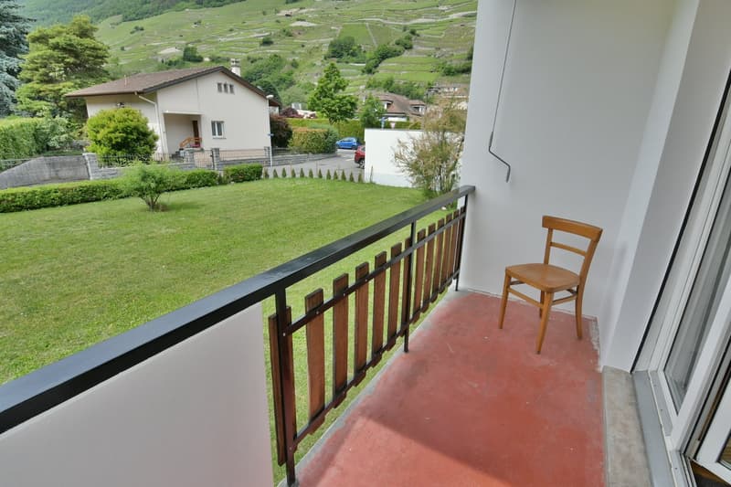 The image shows a balcony or terrace with a wooden railing overlooking a scenic landscape. The balcony has a tiled floor and a wooden chair is visible. The surrounding area includes a residential building, green hills, and trees.