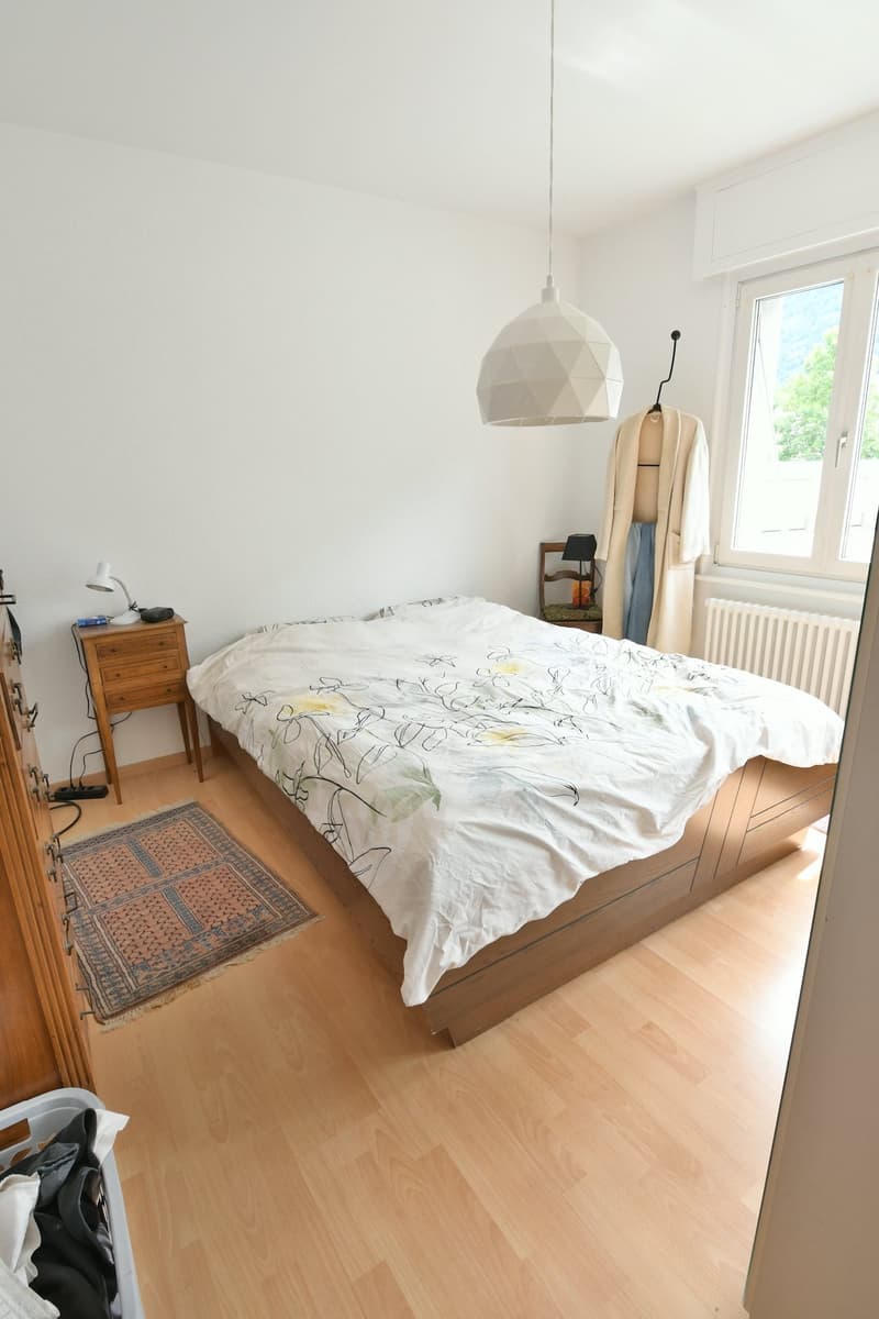Spacious bedroom with hardwood flooring, white walls, a large bed with patterned bedding, a wooden dresser, and a hanging light fixture. There is also a rug on the floor and a window that suggests the presence of a balcony or terrace.