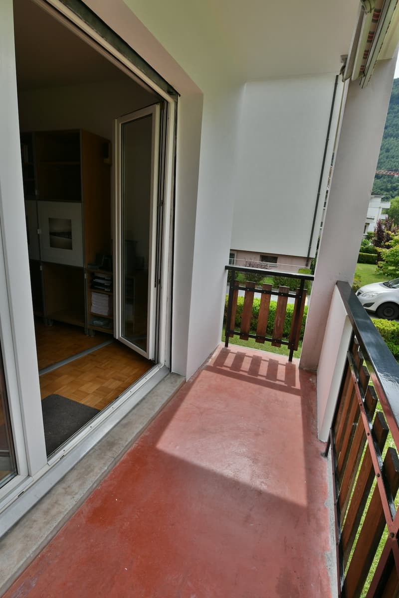 The image shows a balcony or terrace area with a sliding glass door leading into the interior of the apartment or home. The balcony has a wooden railing and the floor is covered in a red tile or concrete material. The view outside the balcony appears to b