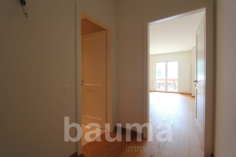 The image shows the interior of an apartment. It has a wooden door leading to another room, and a large window that provides natural light. The floors appear to be hardwood. The walls are painted a neutral color.