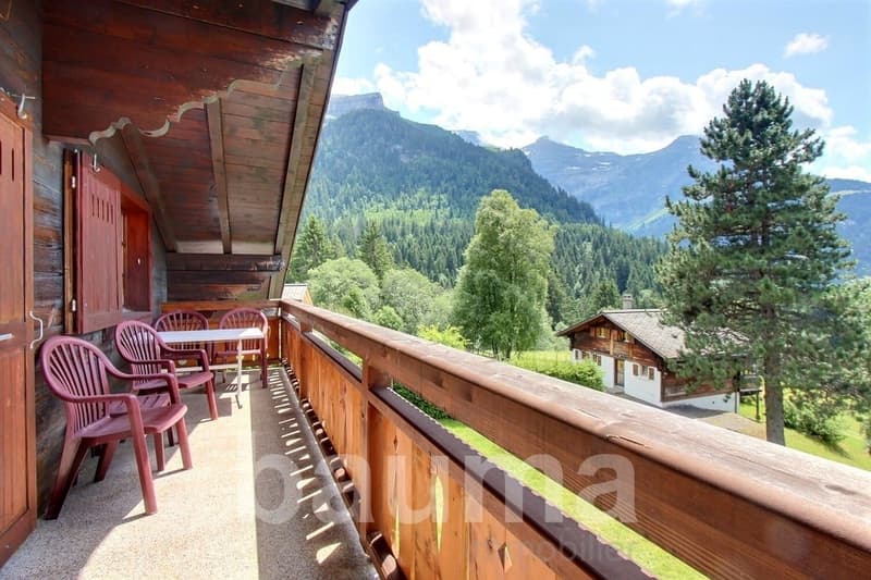 Wooden chalet-style house with a balcony overlooking a scenic mountain landscape with forests and hills. The balcony has wooden chairs and tables, providing a comfortable outdoor seating area to enjoy the beautiful natural surroundings.