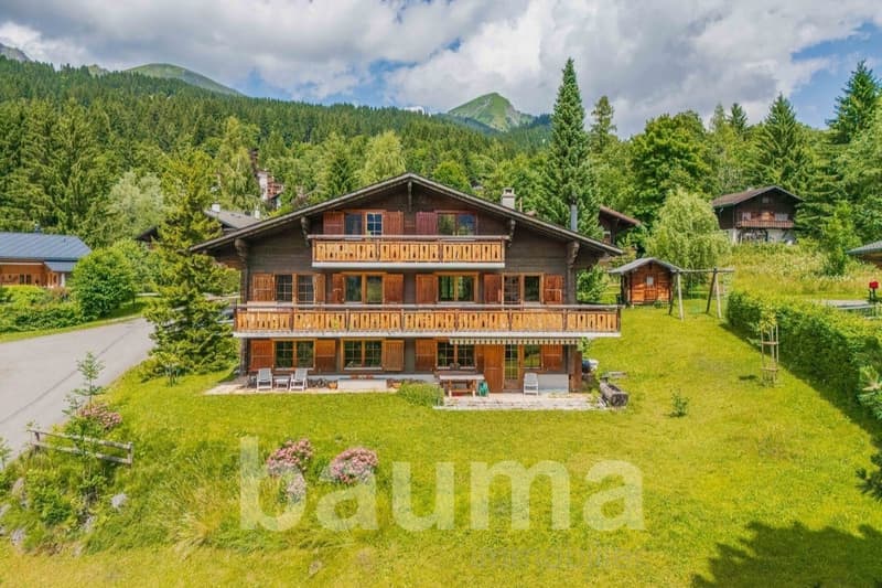 2-story wooden chalet-style house with balconies, surrounded by lush green trees and mountains in the background