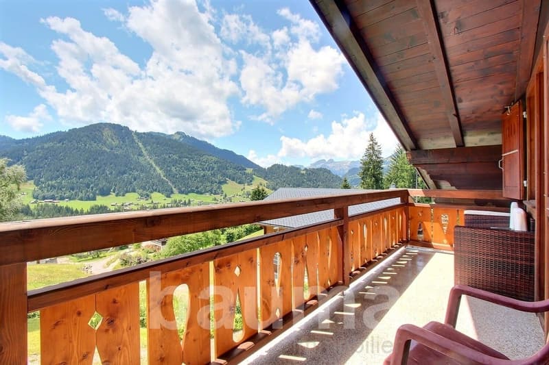 Wooden balcony with a scenic view of the mountains and forests in the distance. The balcony has a wooden railing and is part of a larger building or chalet.