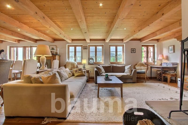 Open space with large windows, wooden beams, beige couches, white rug, coffee table, armchairs, lamp, desk, shelves, picture frames