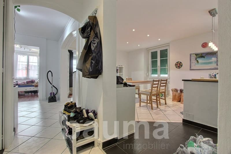 Entrance hall with white tiles, white walls, wooden table, chairs, white shelf with shoes, white ceiling, white door, white cabinet, picture frame, lamp, and a bedroom with a bed and window.