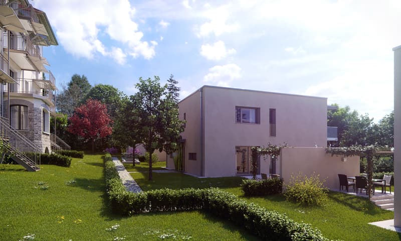 Multi-story apartment building with balconies, surrounded by a well-maintained garden with trees, shrubs, and a paved walkway. There appears to be a parking area or driveway adjacent to the building.