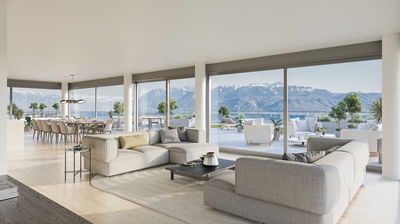Spacious open-plan living area with large windows offering panoramic views of the mountains and lake. The room features a comfortable seating area with grey sofas and armchairs, a coffee table, and a dining area with a long wooden table and chairs. The sp