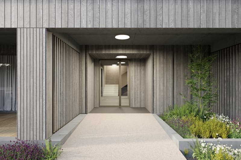 The image shows the entrance of a building with a raised ground floor. The exterior is made of wooden panels and the entrance is framed by a concrete structure. There is a paved walkway leading to the entrance, and the surrounding area is landscaped with