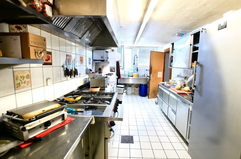 Commercial kitchen with stainless steel appliances, tiled floor, gas stove, oven, sink, refrigerator, and various cooking utensils and ingredients.