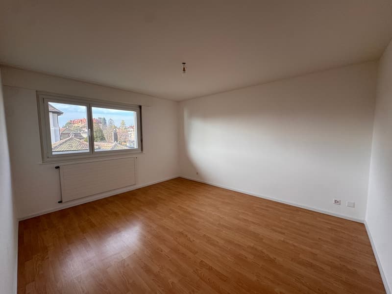 A bare room with white walls, wooden floor, large window with city view, white radiator