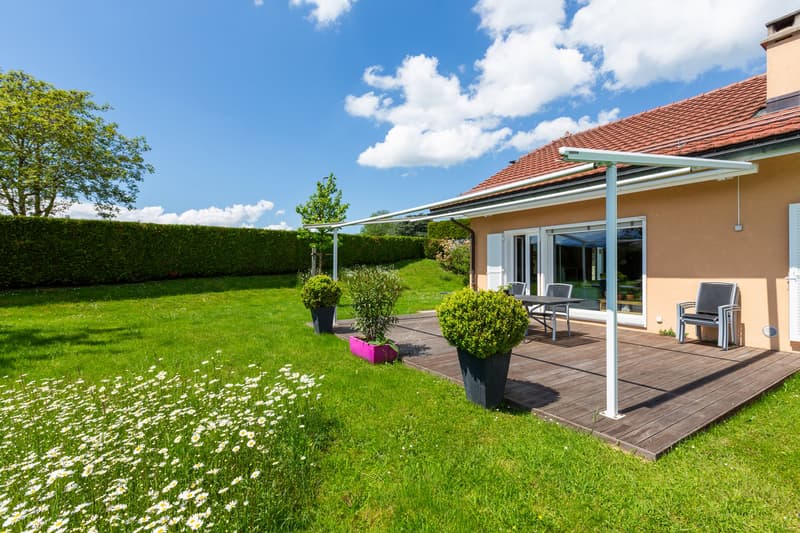 Wooden deck with canopy, outdoor dining table and chairs, lawn with flowers, house with glass doors