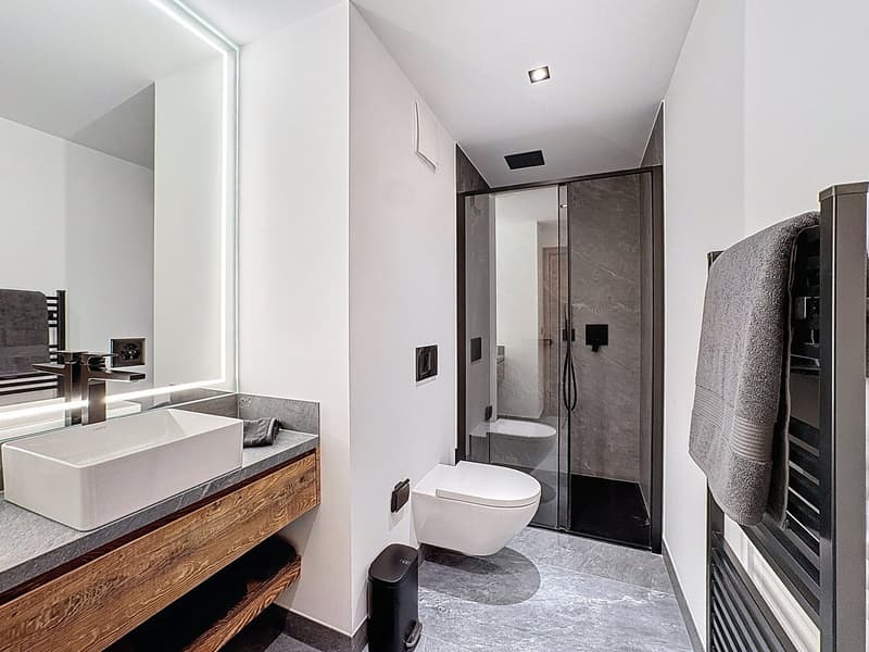 Minimalist bathroom, under-mount sink, wall-mounted toilet, walk-in shower, grey marble flooring, towel rack, grey mirror, wood vanity