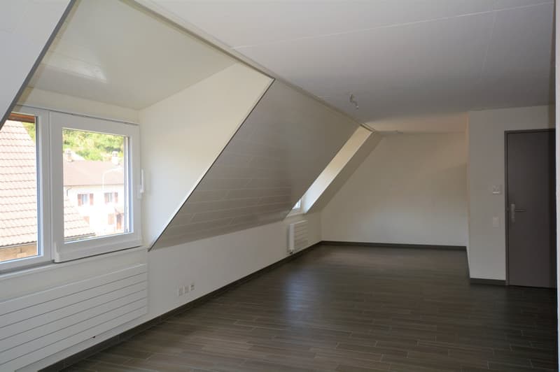 Spacious attic room with slanted ceilings, large window, hardwood-style flooring, and a radiator on the wall.