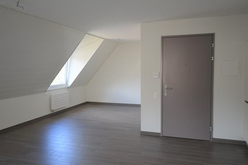Attic room with slanted ceiling, hardwood flooring, and a large window. The room has a plain white wall and a dark brown door.